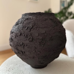 May include: A dark brown, textured ceramic vase with a rounded body and a slightly flared rim. The vase has a rough, organic surface, and sits on a light-coloured, round surface. The overall aesthetic is modern and minimalist.