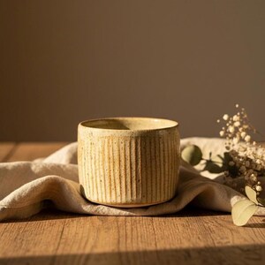 May include: A cylindrical, beige ceramic cup with vertical ribbed detailing. The cup sits on a beige linen cloth atop a wooden surface, with soft lighting and a neutral background. Dried flowers and greenery are in the background.