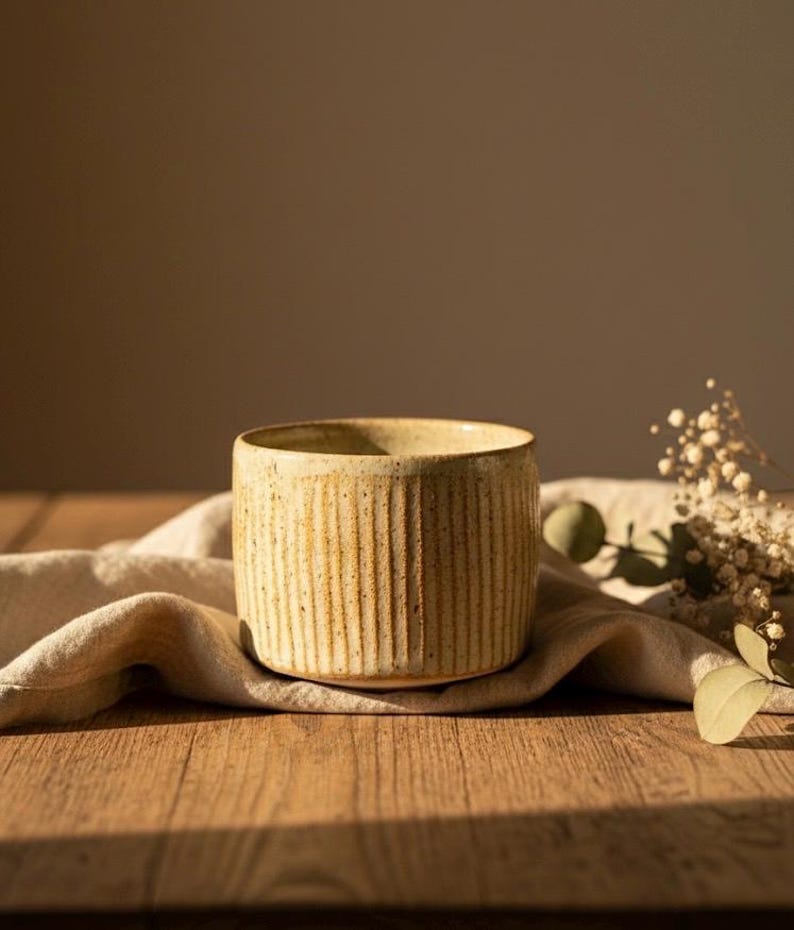 May include: A cylindrical, beige ceramic cup with vertical ribbed detailing. The cup sits on a beige linen cloth atop a wooden surface, with soft lighting and a neutral background. Dried flowers and greenery are in the background.