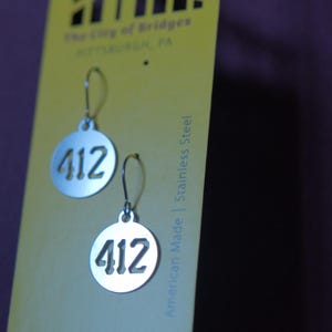 May include: Silver-colored earrings with a circular design, featuring the number "412" cut out in the center. The earrings hang from a simple hook. The background is a yellow card with text that reads "The City of Bridges PITTSBURGH, PA".