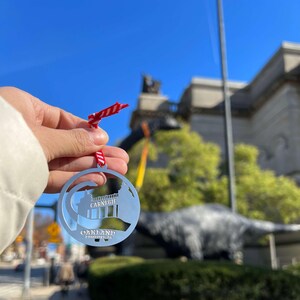 May include: Silver ornament featuring a silhouette of the Carnegie Museum of Natural History in Oakland, Pennsylvania. The ornament is hanging from a red and white striped ribbon.