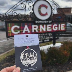 May include: A silver stainless steel ornament featuring a cut-out design of the Carnegie Borough skyline. The ornament is packaged on a black card with the text "Stainless Steel Ornament", "Audra Azoury", "Pittsburgh, PA", "Audra Azoury", "Borough of Carnegie", and "Made in Pittsburgh, PA www.AudraAzoury.com".