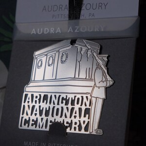 May include: Silver metal ornament depicting a soldier standing guard in front of a building with the words "Arlington National Cemetery" inscribed below. The ornament is made by Audra Azoury.