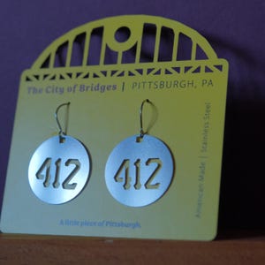 May include: Silver-colored, circular earrings with the number "412" cut out, hanging from a yellow card. The card reads "The City of Bridges | PITTSBURGH, PA" and "A little piece of Pittsburgh." The earrings are made of stainless steel.