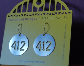May include: Silver-colored, circular earrings with the number "412" cut out, hanging from a yellow card. The card reads "The City of Bridges | PITTSBURGH, PA" and "A little piece of Pittsburgh." The earrings are made of stainless steel.