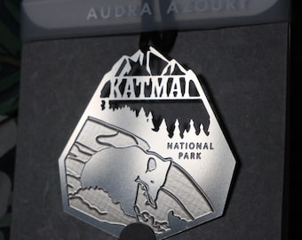 Può includere: Un ornamento in metallo argentato a forma di montagna con le parole "Katmai National Park" incise su di esso. L'ornamento presenta un orso e alberi.
