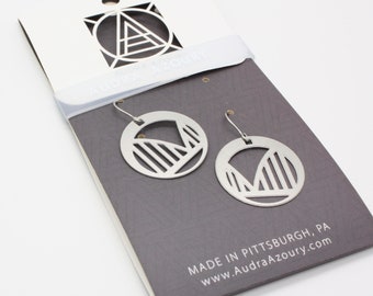 May include: Silver earrings with a geometric design, featuring a circle with a cut-out shape resembling a mountain range. The earrings are displayed on a gray card with the text "Audra Azoury" and "Made in Pittsburgh, PA www.AudraAzoury.com".
