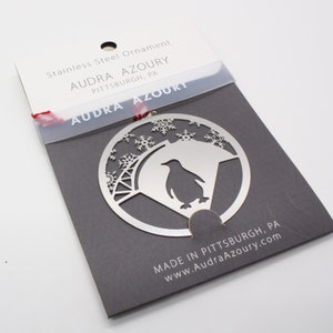 Puede incluir: Un adorno navideño de acero inoxidable plateado con un pingüino y copos de nieve. El adorno está empaquetado en una tarjeta gris con el texto "AUDRA AZOURY PITTSBURGH, PA" y "MADE IN PITTSBURGH, PA www.AudraAzoury.com".