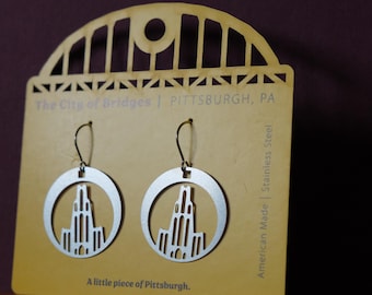 May include: Silver-colored stainless steel earrings featuring a Pittsburgh skyline design within a circular frame. The earrings hang from a wire hook. The card behind the earrings reads "The City of Bridges | PITTSBURGH, PA" and "A little piece of Pittsburgh."
