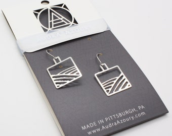 May include: Silver-toned square earrings with a landscape design, displayed on a gray card with the brand name and logo. The earrings have a hook closure. The card reads "MADE IN PITTSBURGH, PA" and the website address.