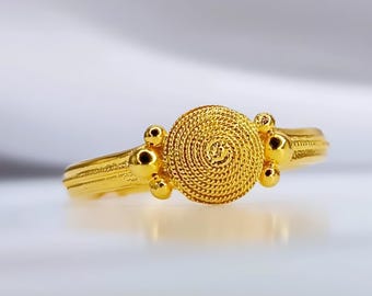 May include: A gold ring with a circular design featuring a spiral pattern. The ring has three small gold balls on each side of the center design.