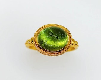 May include: A gold ring with a large, oval green gemstone. The gemstone is set in a gold bezel with a rope-like border. The ring's band is gold, with decorative elements on the sides. The ring is a piece of jewellery.