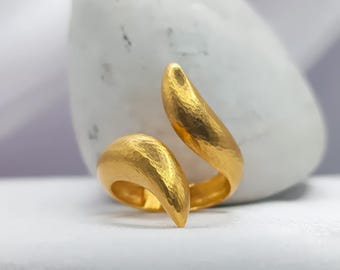 Puede incluir: Un anillo dorado con textura martillada. El anillo tiene un diseño abierto único con dos extremos curvos y puntiagudos que se superponen. El anillo está sobre un fondo blanco.
