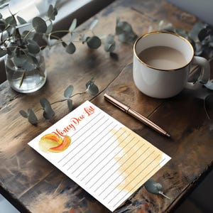 May include: A notepad with the words "Honey Dew List" in red script, featuring a watercolor illustration of a honeydew melon. The notepad has lined pages and a watercolor wash. A rose gold pen and a white mug with a gold rim are also on the wooden surface.