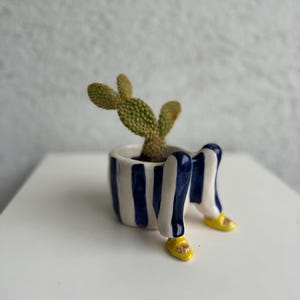 Op de afbeelding: Een grillige keramische plantenpot met een cactus. De pot is wit met marineblauwe verticale strepen en benen met gele sandalen. De cactus heeft groene, getextureerde bladeren. Een uniek decoratief item.
