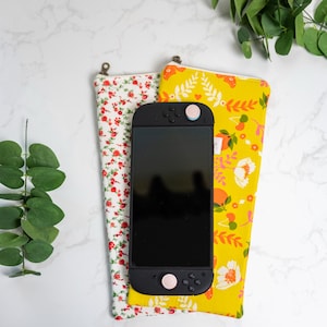May include: A black handheld gaming console rests on top of two fabric cases. One case is white with a red floral pattern, and the other is yellow with a floral design. The console has pink accents.