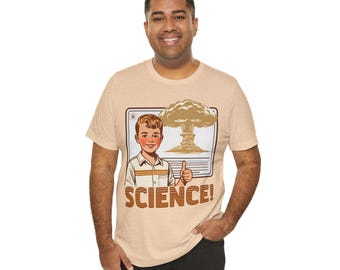 Op de afbeelding: Beige T-shirt met een retro illustratie van een jongen die een duim omhoog steekt, met een paddenstoelwolk op de achtergrond. Het woord "SCIENCE!" is in oranje gedrukt. Het T-shirt heeft een ronde hals en korte mouwen. Vintage design.