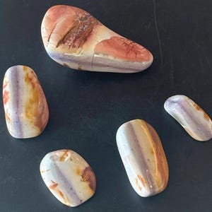 May include: Five polished stones in shades of purple, orange, and cream. The stones have a smooth, rounded shape and a marbled appearance. The largest stone is elongated, while the others are smaller and more oval. The stones are arranged on a dark surface.