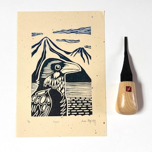 Puede incluir: Un linograbado de un cuervo con montañas de fondo, en tonos azules y negros sobre papel beige. Una herramienta de tallado de madera con una cuchilla negra y mango de madera está junto a la impresión.