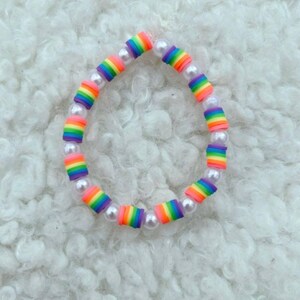 May include: A rainbow-colored beaded bracelet with white pearl beads. The bracelet features small, cylindrical beads in a rainbow pattern of red, orange, yellow, green, blue, and purple. The bracelet is on a white, fluffy surface.