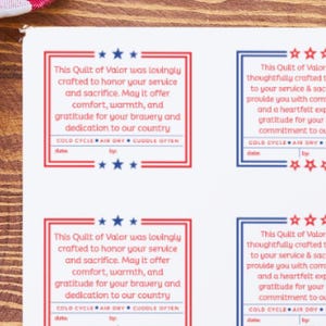 May include: A collection of rectangular labels with patriotic themes, featuring text honoring service and sacrifice. The labels are in red and blue frames with star accents. A rotary cutter and a red and white checkered cloth are also visible.