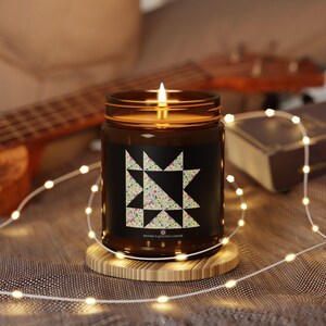 Puede incluir: Un tarro de vela de vidrio marrón con una etiqueta negra que presenta un patrón geométrico colorido. La etiqueta también incluye el texto "BRINDLE'S QUILTING CORNER". La vela está encendida y se encuentra sobre un posavasos de madera.