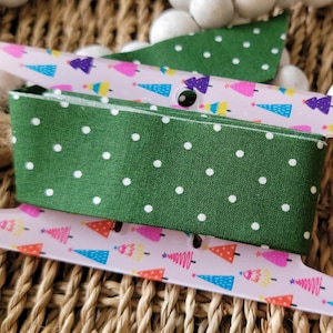 May include: A spool of green fabric with white polka dots is wrapped around a clear plastic spool holder. The holder has a pink and white design with colorful Christmas trees.