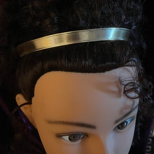 May include: A silver headband with a smooth, shiny finish. The headband is worn on a mannequin with dark brown curly hair.