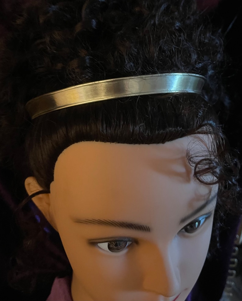 May include: A silver headband with a smooth, shiny finish. The headband is worn on a mannequin with dark brown curly hair.