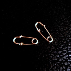 May include: Two gold safety pins on a black background. The safety pins are small and delicate.