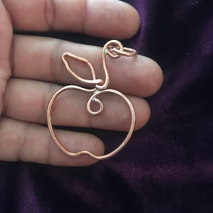 May include: Copper wire apple pendant with a leaf and loop for hanging. The apple design is a continuous line, creating a minimalist and artistic look. The pendant is a warm copper colour, and the design is simple and elegant.