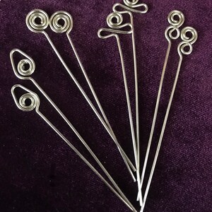 Puede incluir: Conjunto de siete pinchos para comida de alambre met&aacute;lico plateado con diferentes formas decorativas en la parte superior. Los pinchos est&aacute;n dispuestos en forma de abanico sobre una superficie de terciopelo morado.