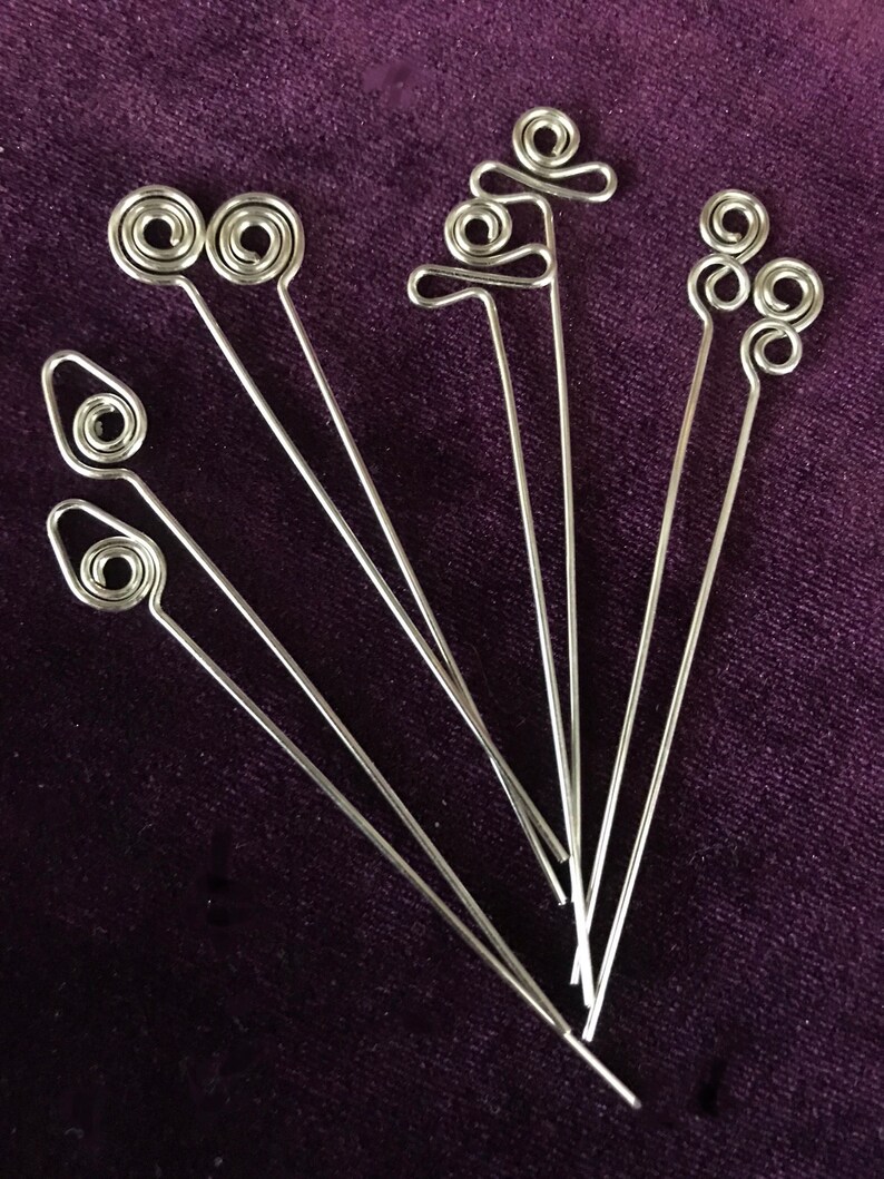 Puede incluir: Conjunto de siete pinchos para comida de alambre met&aacute;lico plateado con diferentes formas decorativas en la parte superior. Los pinchos est&aacute;n dispuestos en forma de abanico sobre una superficie de terciopelo morado.