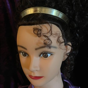 May include: A silver headband with a smooth, shiny finish. The headband is worn on a mannequin with curly brown hair.