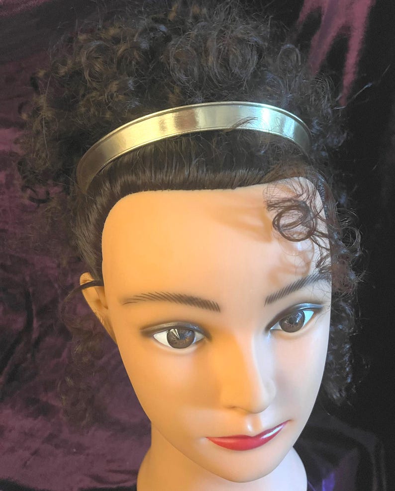 May include: A silver headband with a smooth, shiny finish. The headband is worn on a mannequin with curly brown hair.