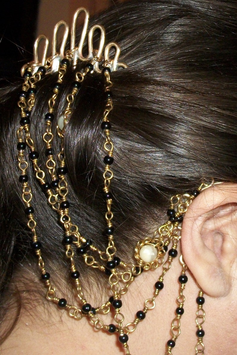 May include: A gold hair comb with black beads and a white pearl-like accent. The comb is worn in a woman's hair, with the beads cascading down the side of her head.