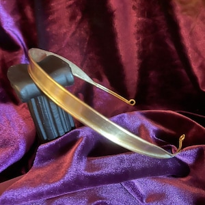 May include: A simple gold-toned metal headband with a smooth, rounded surface. The headband is displayed on a purple velvet background.
