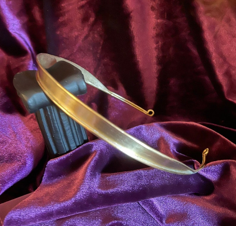 May include: A simple gold-toned metal headband with a smooth, rounded surface. The headband is displayed on a purple velvet background.