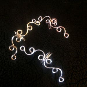 May include: A pair of ear cuffs made of silver and rose gold wire. The cuffs are shaped like a series of swirls and loops.