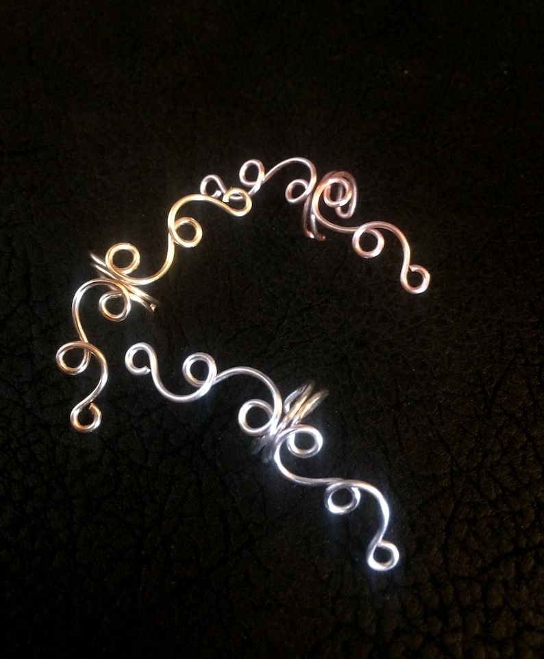 May include: A pair of ear cuffs made of silver and rose gold wire. The cuffs are shaped like a series of swirls and loops.
