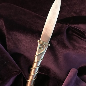 May include: A silver knife with a twisted handle and a gold accent. The knife is lying on a dark purple velvet surface.