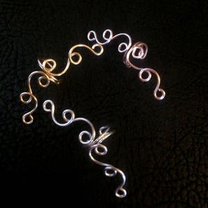 May include: Two gold ear cuffs with a delicate, swirling design. The cuffs are made of thin wire and have a delicate, feminine look.