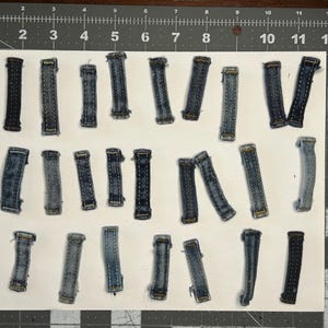 Może przedstawiać: Kolekcja prostokątnych kawałków materiału dżinsowego w różnych odcieniach niebieskiego. Ułożone na białej powierzchni, ukazują fakturę i detale szwów materiału dżinsowego.