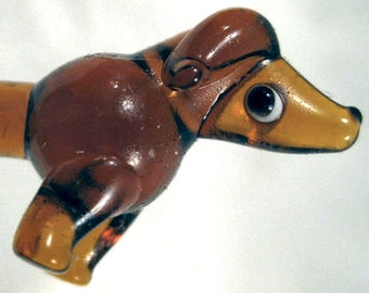 May include: A brown and amber glass figurine of a dachshund dog. The dog has a detailed head, with a black eye and a long snout. The body is brown with black stripes. The figurine is a decorative item.
