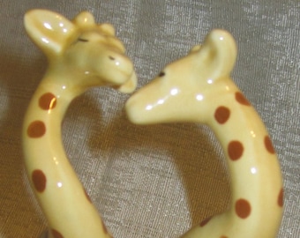 May include: A pair of ceramic giraffe figurines. The giraffes have tan bodies with brown spots, long necks, and black hooves. They are posed as if they are kissing. The figurines are likely salt and pepper shakers.