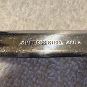 May include: A stainless steel knife blade with a brown handle. The metal surface features visible scratches and is clearly stamped with the text STAINLESS STEEL KOREA near the bolster where the blade meets the handle.