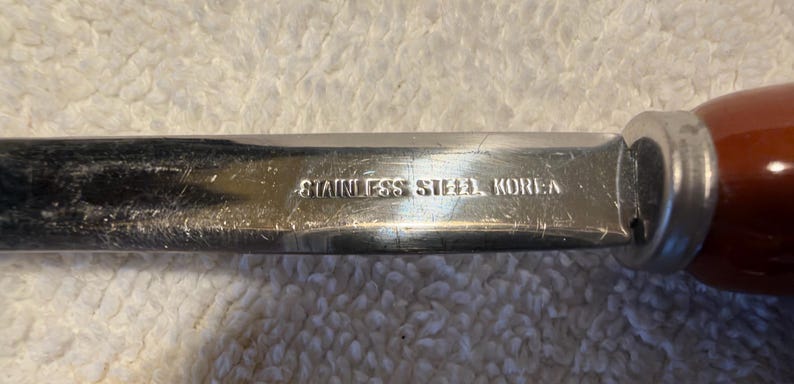May include: A stainless steel knife blade with a brown handle. The metal surface features visible scratches and is clearly stamped with the text STAINLESS STEEL KOREA near the bolster where the blade meets the handle.