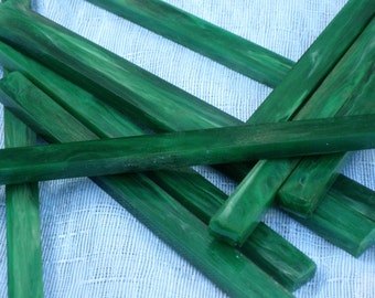 May include: A collection of rectangular, dark green wooden pieces with a glossy finish. The wood grain is visible, and the pieces are arranged in a scattered pattern against a light blue fabric background. The pieces appear to be approximately 1 inch wide.
