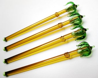 May include: Four glass wands with a yellow-amber hue. Each wand features a green floral design at the top and a green tip. The wands are arranged diagonally against a white background, showcasing their elegant design.