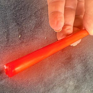 Puede incluir: Una varilla larga, cilíndrica y de color naranja translúcido con una superficie lisa y brillante. La varilla es recta y de grosor uniforme, con un extremo redondeado y un color naranja vibrante que permite el paso de la luz.